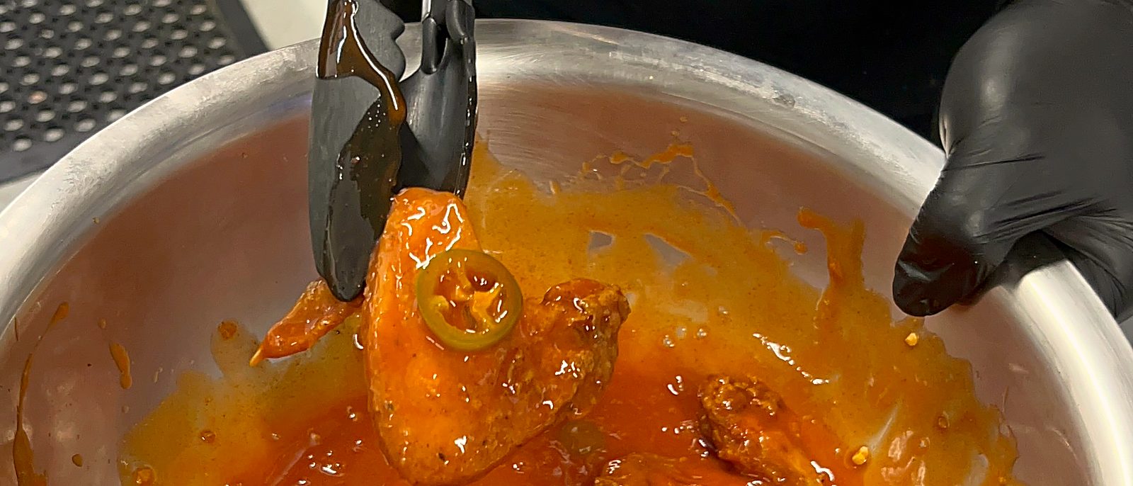 Buffalo-sauced wing with a jalapeño slice lifted from the sauce bowl with tongs